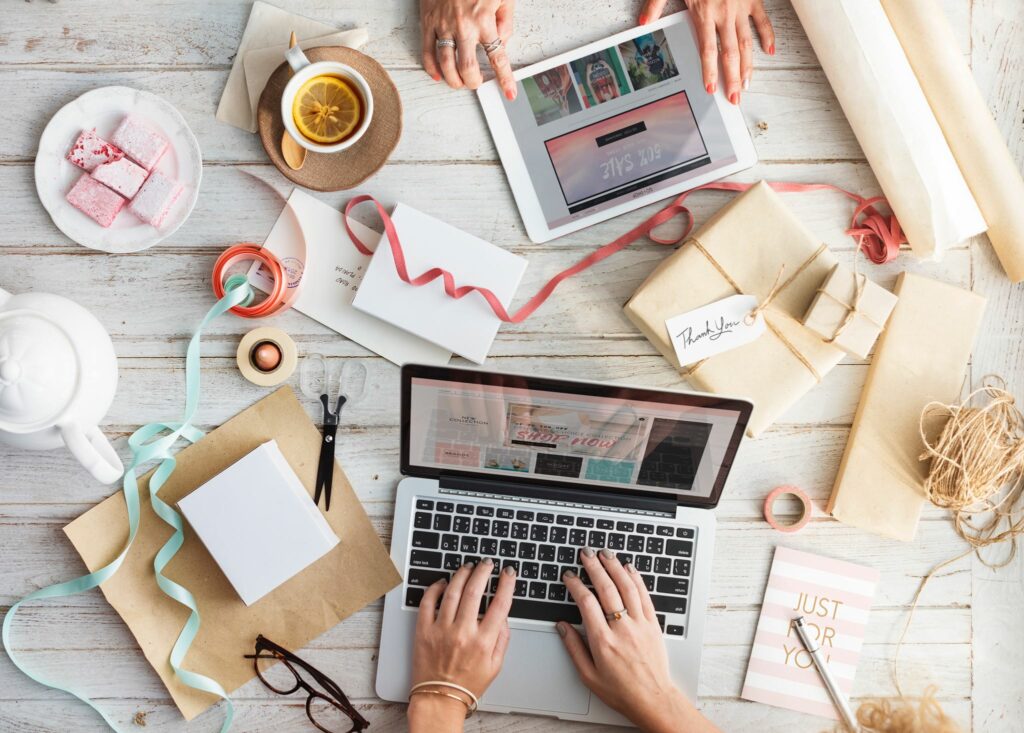 An overhead view of two people working at a table cluttered with packages, twine, paper, and tea.